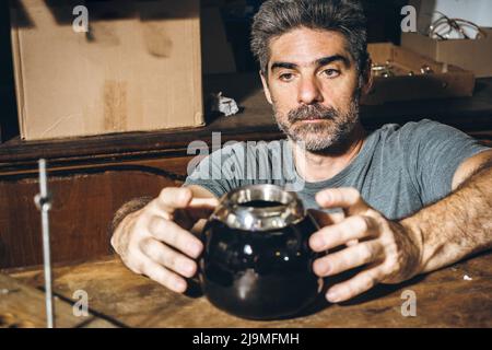 Medio invecchiato bearded maschio artigiano mettendo bordo d'argento fatto a mano su tazza di ceramica nera posto sul tavolo mentre lavora in officina Foto Stock