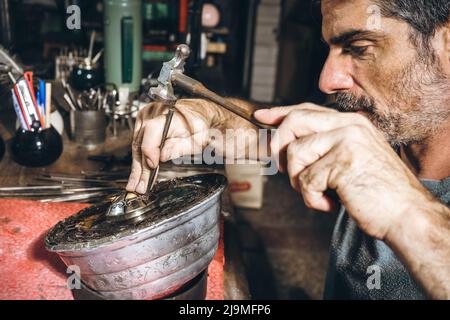 Raccolto uomo maturo bearded con martello e punzone per carve ornamento su argento dettaglio durante il lavoro in officina professionale Foto Stock