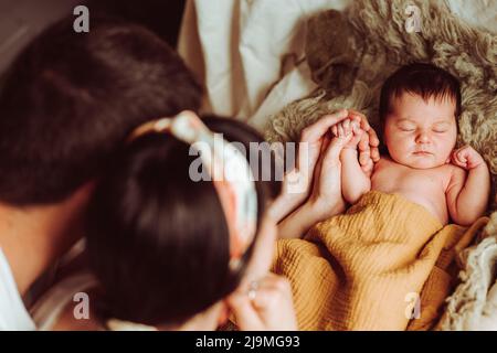 Alto angolo di giovane madre e padre irriconoscibile con capelli scuri che tengono la mano di adorabile neonato che giace e dorme su un letto morbido alla luce del giorno Foto Stock