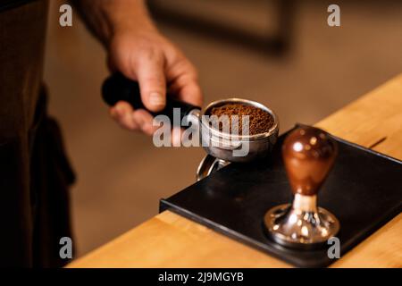Coltivare un barista irriconoscibile che tiene il portafilter con caffè macinato e manomissioni mentre si prepara l'espresso al banco nel moderno caffè Foto Stock