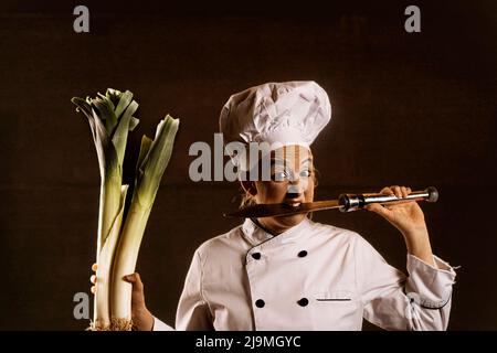 Divertente giovane comico femminile con trucco in costume da chef e cappello mordente coltello affilato e tenendo porri di palude su sfondo nero Foto Stock