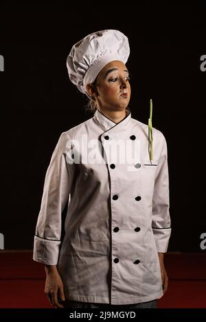 Giovane artista mimo femminile con trucco divertente in abiti chef e cappello fare il viso e guardare lontano su sfondo nero Foto Stock