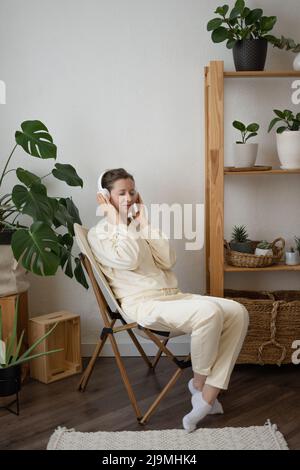 Tutta la lunghezza di giovane donna rilassata in abiti casual seduti su sedia e ascoltare musica con occhi chiusi in cuffie wireless in un appartamento chiaro Foto Stock