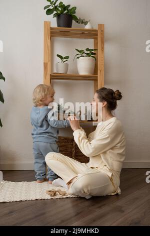 Corpo pieno di felice giovane madre nufooted che gioca con adorabile ragazzino in abiti casual seduti sul pavimento in parquet a casa Foto Stock