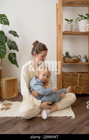 Corpo pieno di felice giovane madre nufooted che tiene e abbraccia adorabile ragazzino in abiti casual seduti sul pavimento in parquet a casa Foto Stock