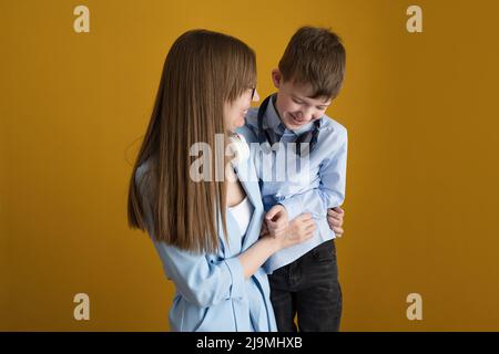 Mamma adulta felice con capelli lunghi che abbraccia e solletico ridacchiando figlio mentre si diverte insieme su sfondo giallo Foto Stock