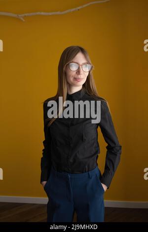 Donna adulta in abiti casual elegante e occhiali guardando lontano mentre si alza contro la parete gialla a casa Foto Stock