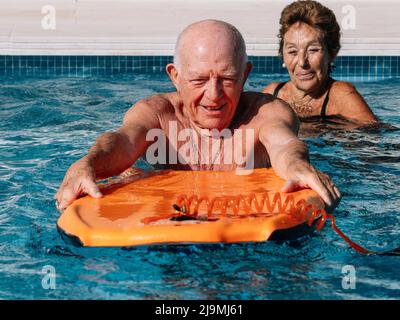 Marito adulto felice che si appoggia sul kickboard di aiuto mentre la moglie nuota nella piscina all'aperto nella giornata di sole Foto Stock