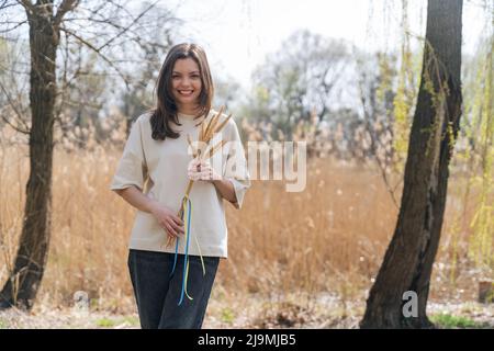 Giovane ragazza allegra con lunghi capelli scuri in abiti casual sorridente e guardando la macchina fotografica mentre in piedi in natura con un mazzo di spikelets di grano legato Foto Stock