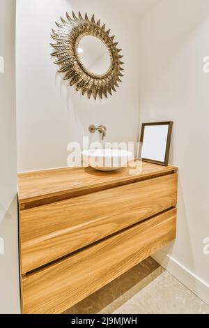 Lavandino in ceramica installato sulla credenza vicino a una cornice vuota sotto un elegante specchio a forma di sole nella moderna stanza da bagno di casa Foto Stock