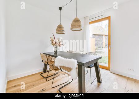 Interni di moderna sala da pranzo in stile minimalista con tavolo nero e sedie in vimini situate sotto le lampade vicino a una porta di vetro durante il giorno Foto Stock