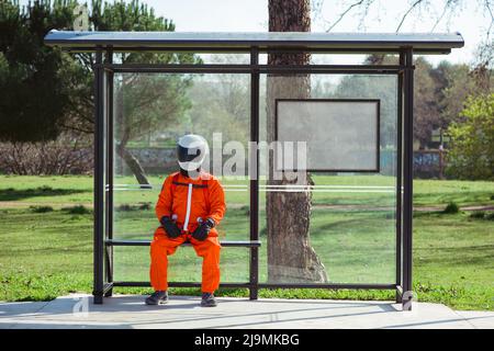 Intera lunghezza di astronauta maschile anonimo in tuta spaziale arancione e casco seduti in panchina alla fermata dell'autobus e in attesa dei trasporti pubblici il giorno di sole Foto Stock