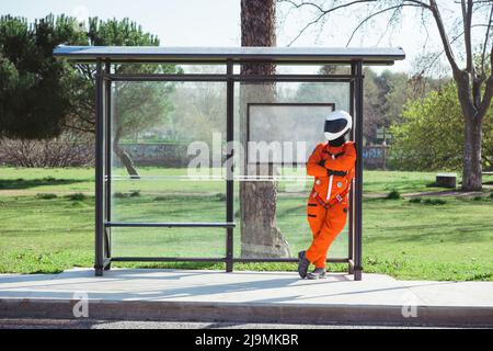 Intera lunghezza di astronauta maschile anonimo in tuta spaziale arancione e casco in piedi alla fermata dell'autobus e in attesa di trasporto pubblico il giorno di sole Foto Stock