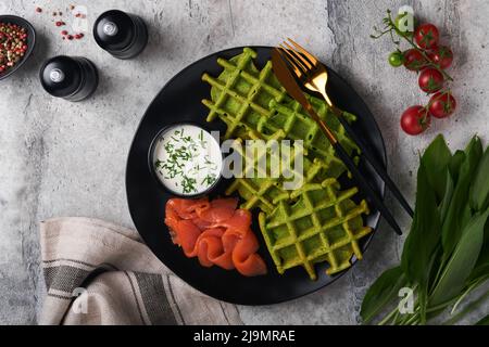 Waffle belgi verdi. Spinaci o aglio selvatico o pesto cialde con salmone rosso e salsa di panna su sfondo grigio tavolo di cemento. Deliziosa colazione, Foto Stock