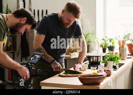 Cameraman professionale video di tiro di uomo caucasico bearded che taglia il prezzemolo fresco su asse di legno Foto Stock
