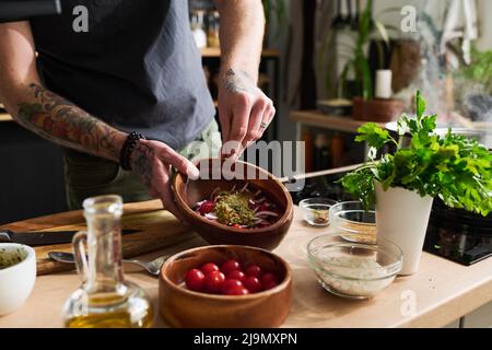 msn non riconoscibile con tatues sulle braccia in piedi al banco cucina cottura insalata di verdure aggiungere sale e pepe in ciotola Foto Stock