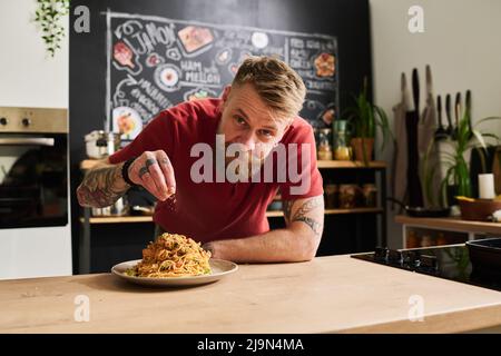 Ritratto dell'uomo caucasico con barba sul viso e tempo libero a casa per finire la cottura della pasta aggiungendo delle spezie sopra di esso Foto Stock