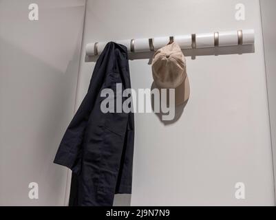 Vista ad angolo basso di una giacca blu navy e di un cappellino da baseball tan appeso a un rack appeso a parete all'interno di un armadio bianco illuminato Foto Stock