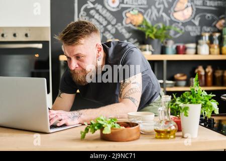 Elegante caucasico bearded uomo cercare ricetta pasto o SMS messaggio in Internet sul portatile in cucina loft a casa Foto Stock