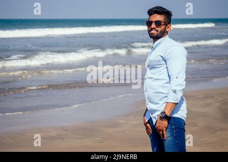 Giovane uomo afro-americano in camicia blu e jeans sulla spiaggia dell'oceano. Foto Stock