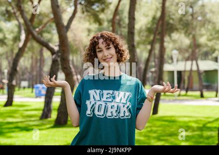 Giovane donna rossa felice indossando casual t-shirt verde in piedi sul parco della città, all'aperto senza indizio e confuso con le braccia aperte, spalle che grattugiano, no Foto Stock