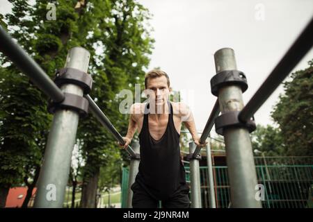 Un ritratto di un uomo muscoloso concentrato in abiti da allenamento neri che fanno salti su barre parallele. Basso angolo di mans fitness in background e spazio aperto intorno a lui. Foto Stock