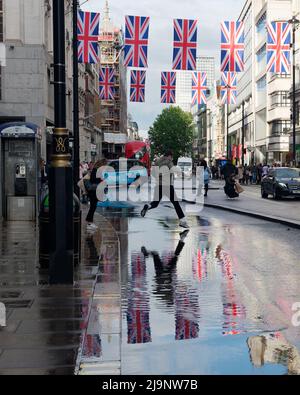 Mans salta attraverso le pozze d'acqua a Oxford Street dopo una pioggia pesante, con le bandiere di Union Jack riflesse nell'acqua. Foto Stock