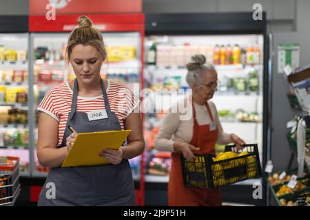 Giovane assistente di negozio in supermercato in guscio di verdure, in pankground è il suo collega riempimento scorte. Foto Stock