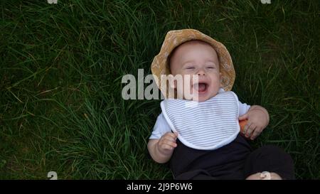 Piccolo bambino neonato felice in estate panama cappello cadere giù posa su erba a piedi nudi in estate Sunny Day. Bambino bambino bambino ragazzo Smacinatura faccia guardare Foto Stock