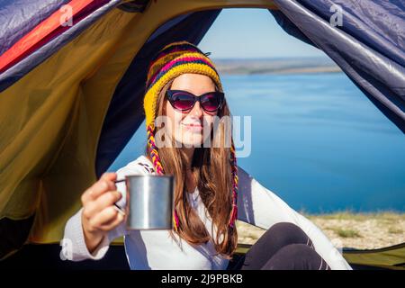 Giovane donna in un divertente cappello dal Nepal seduta in un sacco a pelo blu nella tenda che tiene una tazza thermos e bere caffè tè sfondo Foto Stock