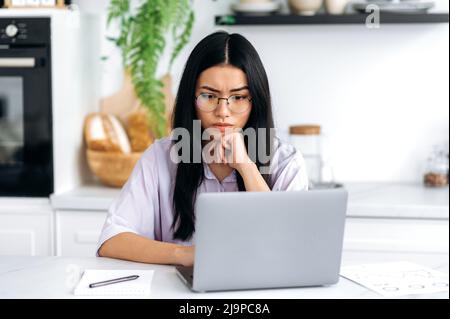 Focalizzata bruna ragazza di nazionalità asiatica con occhiali, freelance, designer, trader, si siede in cucina, guarda concentrato al laptop, lavora a un importante progetto, analizza i risultati Foto Stock