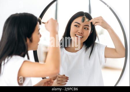 Felice bruna cinese bella ragazza in t-shirt bianca, si alza di fronte a uno specchio a casa, fa con attenzione la sua routine di bellezza mattina, si prende cura della sua pelle facciale, utilizza un rullo di pietra facciale, sorridente Foto Stock