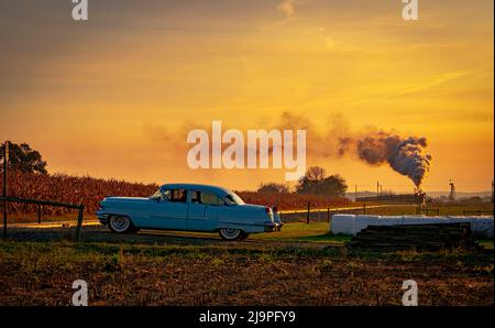 Una vista di un treno passeggeri a vapore antico che si avvicina all'alba con una testa piena di vapore e fumo che viaggia attraverso le terre con un'auto d'epoca in attesa di passare Foto Stock