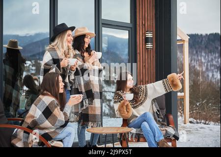 Le giovani donne che si godono week-end invernali sulla terrazza della casa contemporanea fienile. Quattro ragazze che si divertono, seduti sulle sedie e bevendo tè caldo. Foto Stock