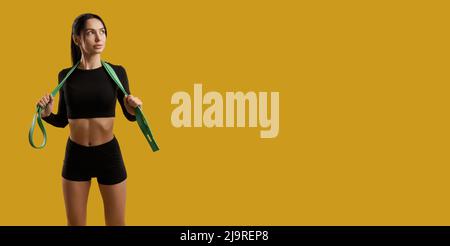 Vista frontale della bella ragazza con ponytail in piedi, allenarsi al chiuso. Donna sottile che indossa la parte superiore nera e gli shorts, guardando da parte, che tiene le fasce di gomma sul collo. Concetto di gioventù. Foto Stock