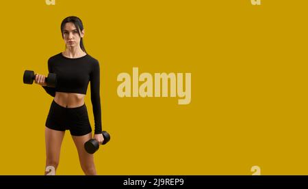 Vista frontale della ragazza piuttosto slim in abbigliamento sportivo che si allenano, si allenano, si allenano. Donna brunetta con ponytail in piedi, sollevando manubri, guardando la macchina fotografica. Concetto di gioventù. Foto Stock