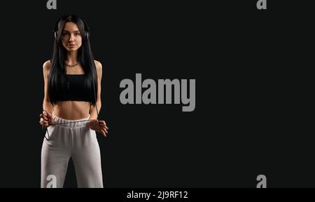 Vista frontale della ragazza brunette con capelli lunghi che ascoltano musica in auricolari. Donna piuttosto sottile che indossa la parte superiore nera e pantaloni bianchi in piedi, guarda la fotocamera ar. Concetto di gioventù. Foto Stock