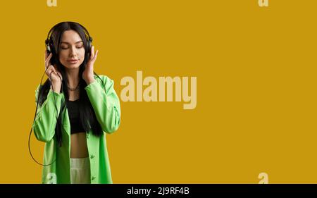 Vista frontale della ragazza indossando tuta verde, in piedi, ascoltando musica. Bella donna bruna con capelli lunghi godendo di musica con occhi chiusi, sorridente. Concetto di vita moderna. Foto Stock