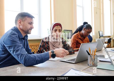 Insegnante caucasico maturo che tiene la penna mostrando qualcosa sullo schermo del portatile e spiegandolo a donna musulmana in hijab durante la lezione Foto Stock