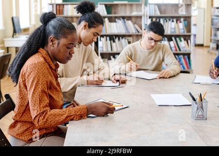 Gruppo di studenti immigrati etnicamente diversi che imparano la lingua inglese seduti in biblioteca facendo test grammaticali Foto Stock