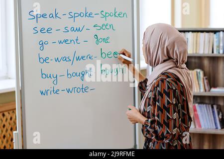 Donna irriconoscibile che indossa il hijab in piedi alla lavagna facendo il compito con verbi irregolari durante la lingua inglese per la lezione immigrati Foto Stock