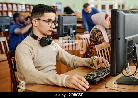 Gruppo di studenti etnicamente diversi seduti in una moderna biblioteca universitaria che lavora con i computer Foto Stock