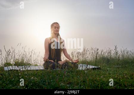 Bella donna con occhi chiusi su tappeto yoga in posa di loto, profondamente respirante e sorridente. Bella donna in tuta sportiva tra erba rilassante dopo la pratica yoga. Concetto di equilibrio. Foto Stock