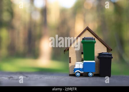 Camion dei rifiuti giocattolo e bidoni dei rifiuti in piedi vicino ad un modello di casa in miniatura all'aperto. Gestione dei rifiuti nelle zone rurali. Foto Stock