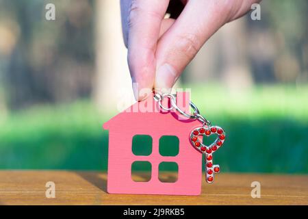 Mano che tiene una casa piatta in legno forma con un piccolo cuore a forma di chiave decorata con rhinestones rosso all'aperto. Campagna concetto di casa. Foto Stock