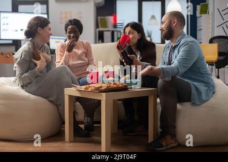 Diversi gruppi di colleghi godono dopo il lavoro festeggiamenti drink in ufficio. Amici allegri e multietnici seduti sul divano con spuntini, patatine e tazze di birra, bevande alcoliche sul tavolo. Foto Stock