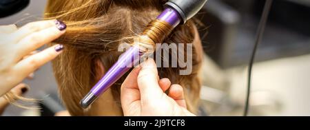 L'acconciatore ricci acconciature di capelli lunghi marrone con il ferro arricciacapelli in parrucchiere, primo piano Foto Stock
