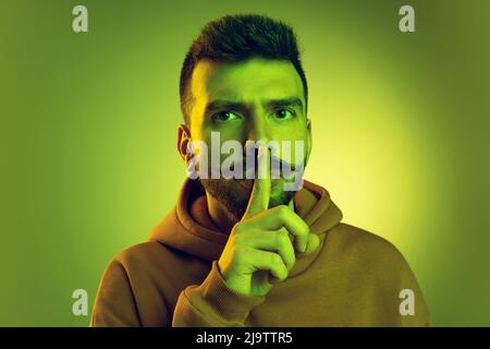 Ritratto di giovane uomo che mette il dito sulle labbra, posa isolata su sfondo verde studio Foto Stock