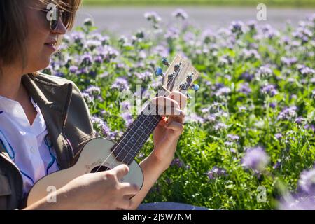 Donna felice che gioca a ukulele in prato fiorito. Divertimento di musica e primavera all'aperto Foto Stock