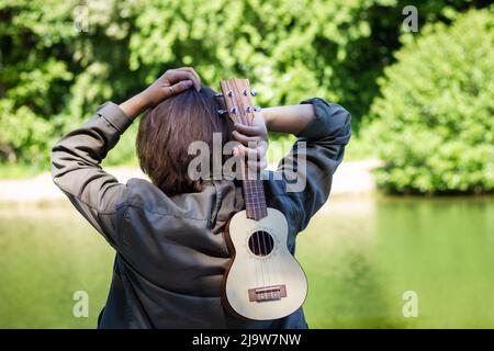 Vista posteriore della donna che tiene l'ukulele all'aperto. Sfondo verde naturale Foto Stock